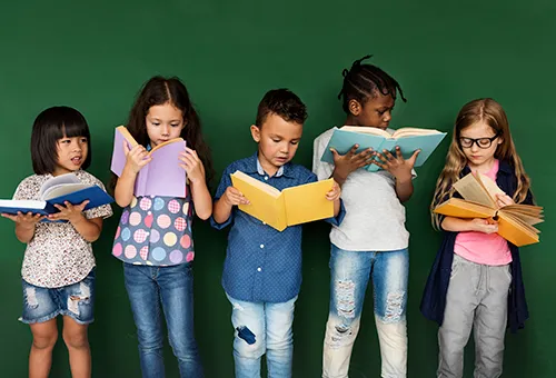Five young children standing in a row against a green wall, each reading an open book